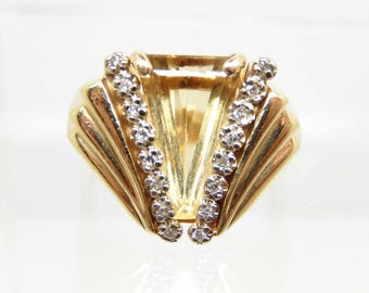 May include: A gold-toned ring with a large, rectangular, light-yellow gemstone at the center. Two rows of small, round, clear gemstones flank the central stone. The ring has a fan-like design on either side of the central stone.