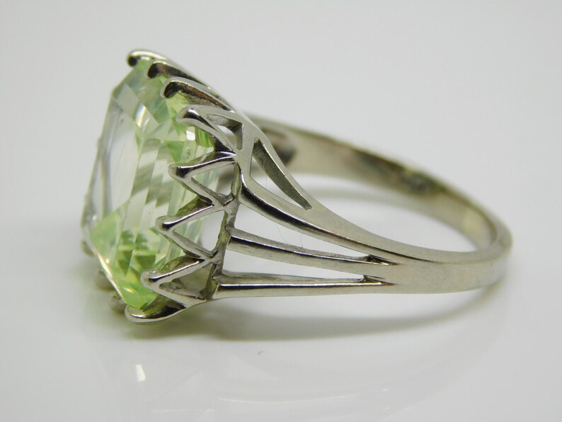 May include: A silver ring with a large, light green, emerald-cut gemstone. The ring has a delicate, openwork band.
