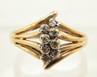 May include: A gold-toned ring with a cluster of small, round diamonds. The ring has a multi-band design with a curved, asymmetrical shape. The diamonds are set in a cluster at the top of the ring.