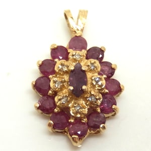 May include: A gold pendant featuring a central marquise-cut ruby surrounded by a cluster of round rubies and small accent diamonds. The floral-shaped design includes a gold bail at the top for attachment to a chain, showcasing a vibrant red color.