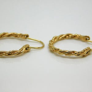 May include: A pair of gold-colored hoop earrings. The earrings have a twisted rope design with small beaded accents. The earrings are displayed on a white surface.