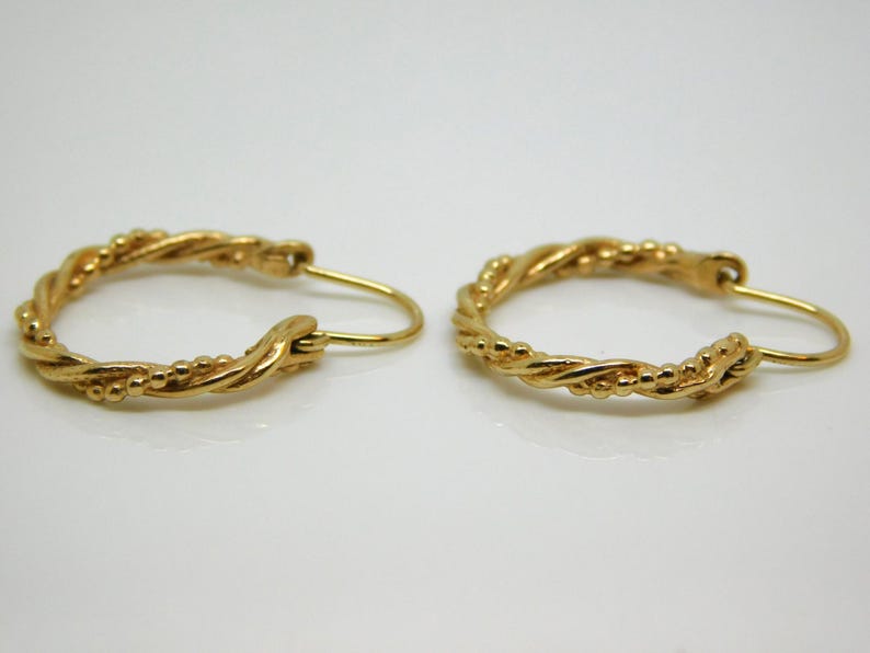 May include: A pair of gold-colored hoop earrings. The earrings have a twisted rope design with small beaded accents. The earrings are displayed on a white surface.