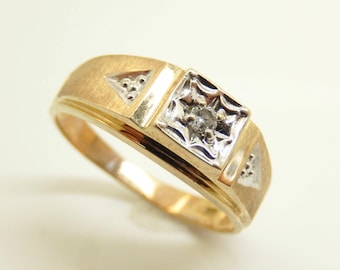 May include: A gold ring with a square, diamond-studded center. The ring has a brushed finish and triangular accents on each side of the center stone. The band is a classic, rounded design.