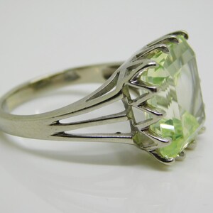 May include: A silver ring with a large, light green, emerald-cut gemstone. The ring has a delicate, openwork design.