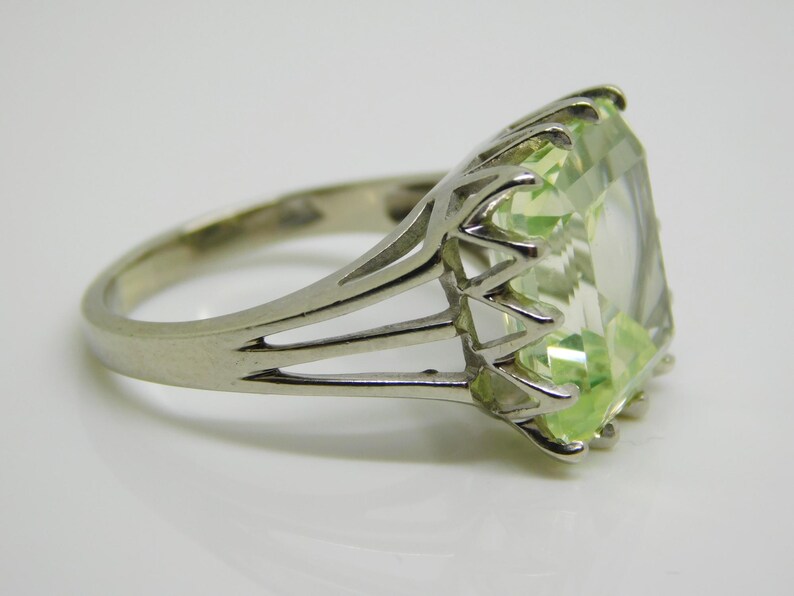 May include: A silver ring with a large, light green, emerald-cut gemstone. The ring has a delicate, openwork design.