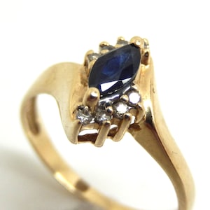 May include: A gold ring featuring a central marquise-cut dark blue sapphire gemstone. The sapphire is flanked by small, clear round-cut stones set into the gold band, which has a smooth, polished finish and a bypass-style design.