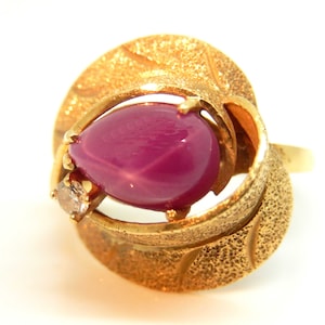 May include: A gold ring with a large pink star-shaped ruby gemstone and a smaller diamond accent. The ring has a unique design with a textured band and a leaf-like setting.