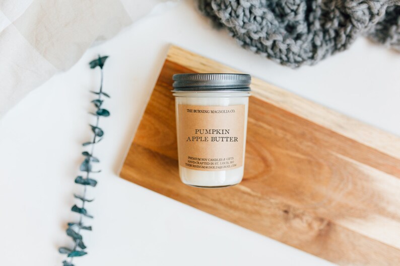Może przedstawiać: Świeca w słoiku z metalową pokrywką i etykietą z napisem "Pumpkin Apple Butter". Świeca stoi na drewnianej desce do krojenia. Tło obejmuje białą powierzchnię, gałązkę zieleni i szary dzianinowy przedmiot.
