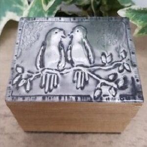 May include: A small wooden box with a decorative lid. The lid features a raised relief design of two birds perched on a branch. The birds are light grey with white accents, and the box has a natural wood finish.