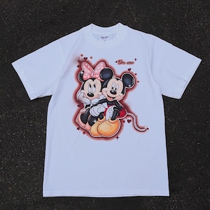 Op de afbeelding: Wit T-shirt met een afbeelding van Mickey en Minnie Mouse die elkaar omarmen. Minnie draagt een roze strik. Het ontwerp bevat de tekst "Be mine". Het shirt heeft korte mouwen en een ronde hals.