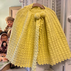 May include: A bright yellow, crocheted cape with a scalloped edge and two pom-poms. The cape is hanging on a wooden hanger. The background includes several vintage dolls.