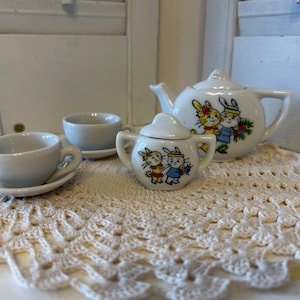 May include: A white porcelain tea set with a teapot, creamer, and two cups. The teapot has a cartoon design of two bunnies with flowers. The tea set is on a white crocheted doily.