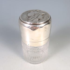 Puede incluir: Un tarro de vidrio con tapa plateada, con un relieve detallado del perfil de una mujer en la tapa. La mitad inferior del tarro es de vidrio transparente grabado, mientras que la parte superior es de metal plateado brillante. El tarro es cilíndrico y está en posición vertical.