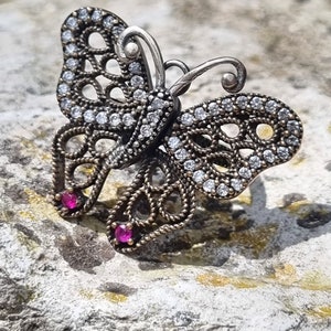 May include: A silver butterfly pendant with clear rhinestones and two pink gemstones. The pendant is on a rough, gray surface.