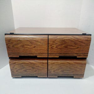 May include: A wooden storage cabinet with four drawers. The cabinet has a dark brown wood grain finish with black trim. The drawers are arranged in two rows of two, with a curved design on the front.