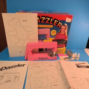 May include: A pink BeDazzler setting machine with a box featuring a girl and the text "AZZLER SETTING MACHINE". Includes pattern sheets with designs for a bear, car, flower, heart, dog, and airplane. Also includes metal studs and glue.