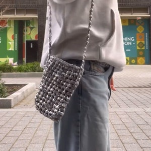 May include: A silver chain strap bag with a woven texture. The bag is worn over the shoulder and is hanging on the side of a person wearing a grey hoodie and blue jeans.