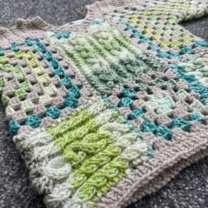 Op de afbeelding: Een handgehaakte trui in een granny square patroon. De trui heeft een mix van kleuren, waaronder lichtgroen, blauwgroen, wit en beige. De mouwen en de halslijn zijn afgewerkt met een effen beige rand. Het is een gezellig, handgemaakt kledingstuk.