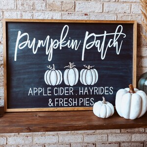 May include: A wooden sign with a chalkboard surface that reads "Pumpkin Patch, Apple Cider, Hayrides & Fresh Pies". The sign is decorated with white painted pumpkins and has a brown wood frame.