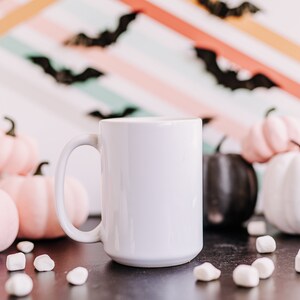 May include: A white ceramic coffee mug with a black background. The mug is surrounded by pink and white pumpkins and white marshmallows.