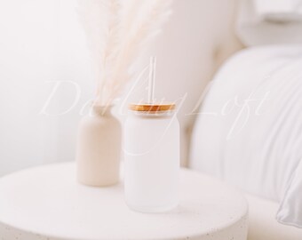 May include: A frosted glass tumbler with a wooden lid and a clear drinking straw. The tumbler is sitting on a white table with a white vase and dried flowers in the background.