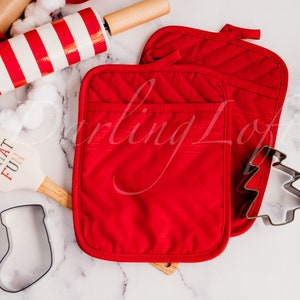 May include: Two red quilted oven mitts with a pocket on the front. The mitts are on a white marble surface with a wooden rolling pin, a white spatula with the text "Oh What Fun", and a metal Christmas tree cookie cutter.