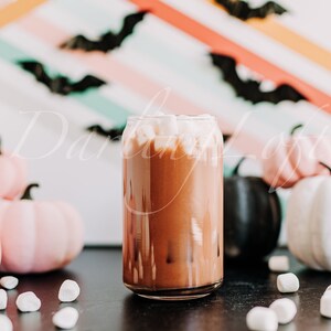 May include: A glass mug filled with hot chocolate topped with marshmallows. The mug is surrounded by pink and white pumpkins and marshmallows on a black surface. The background is a colorful striped wall with black bats.