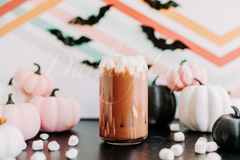 May include: A glass mug filled with hot chocolate topped with marshmallows. The mug is surrounded by pink and white pumpkins and marshmallows on a black surface. The background is a colourful striped wall with black bats.