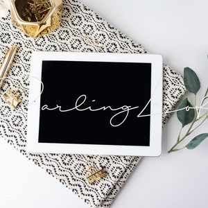May include: A white tablet computer with a black screen displaying the text "Darling Loft" in white cursive script. The tablet is lying on a black and white patterned throw blanket with a gold pen and a gold paperclip. A sprig of eucalyptus leaves is on the right side of the image.