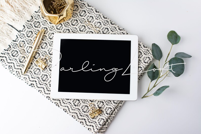 May include: A white tablet computer with a black screen displaying the text "Darling Loft" in white cursive script. The tablet is lying on a black and white patterned throw blanket with a gold pen and a gold paperclip. A sprig of eucalyptus leaves is on the right side of the image.