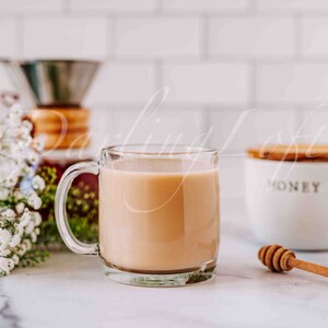 May include: A glass mug filled with a light brown beverage, such as coffee or tea, sits on a white countertop. A small white container with a wooden lid and the word "honey" printed on it is to the right of the mug. A wooden honey dipper is lying on the counter.