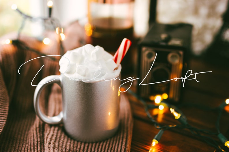 May include: A silver mug filled with hot chocolate topped with whipped cream and a candy cane. The mug is sitting on a brown knitted blanket with string lights in the background.
