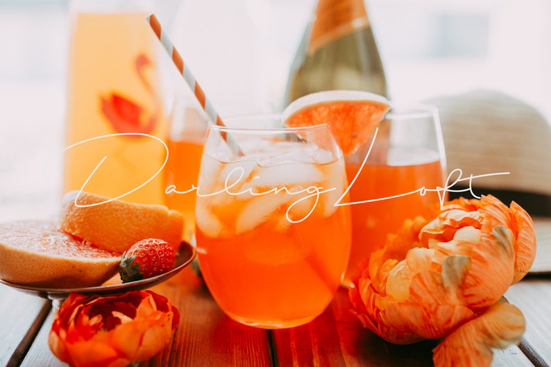 Puede incluir: Dos vasos de zumo de naranja con hielo y una rodaja de pomelo como guarnici&oacute;n. Una botella de champ&aacute;n y una jarra de zumo de naranja est&aacute;n en el fondo. Los vasos est&aacute;n sobre una mesa de madera con flores naranjas y una rodaja de pomelo.