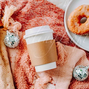 May include: A white paper coffee cup with a brown sleeve sits on a textured, peach-colored blanket, next to a plate of glazed donuts. Two disco ball ornaments add a festive touch to the scene.