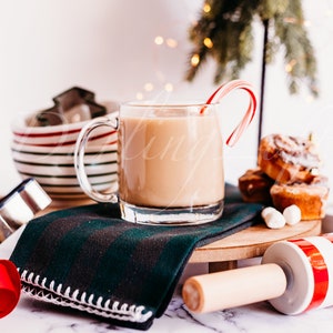 May include: A mug of hot chocolate with a candy cane stirring stick, a green and white striped bowl, a metal cookie cutter, a red ribbon, a green and black plaid napkin, a wooden rolling pin with red and white stripes, and cinnamon rolls on a wooden tray.