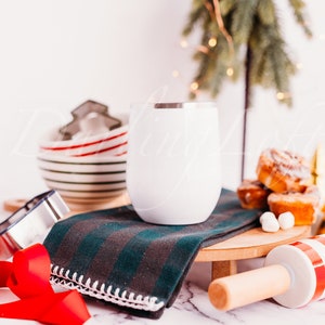 May include: A white stainless steel wine tumbler with a silver rim sits on a green and black striped kitchen towel. The tumbler is on a wooden cutting board with a red and white striped rolling pin and a bowl of cinnamon rolls.