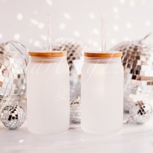 May include: Two frosted glass tumblers with bamboo lids and clear straws. The tumblers are sitting on a white surface with silver disco balls in the background.