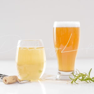 May include: Two glasses of pale yellow liquid, one in a tall glass and one in a round glass. A sprig of green rosemary sits on a white surface next to the glasses.
