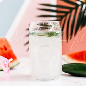 May include: A glass of lemonade with ice and a lime wedge, next to a slice of watermelon and a green jalapeno pepper. The drink is in a clear glass with a white background.