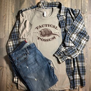 Puede incluir: Una camiseta de color beige con un gr&aacute;fico marr&oacute;n de un armadillo con el texto "Tactical Possum" en letras marrones.