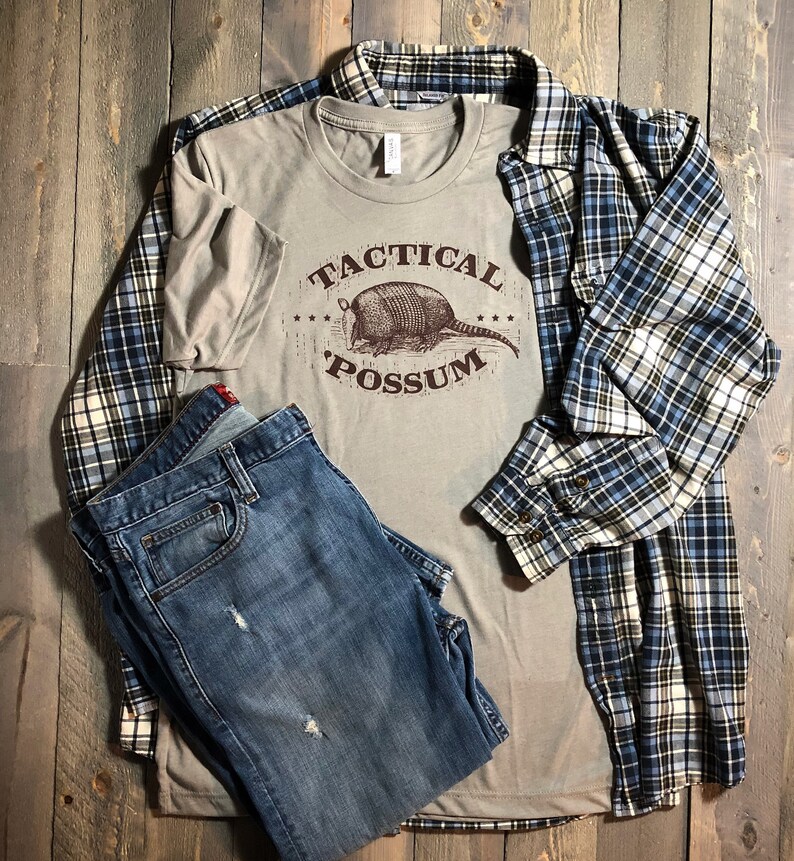Puede incluir: Una camiseta de color beige con un gr&aacute;fico marr&oacute;n de un armadillo con el texto "Tactical Possum" en letras marrones.