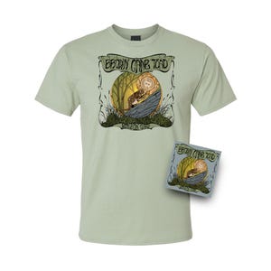 Op de afbeelding: Lichtgroen t-shirt met een afbeelding van een bruine pad die een Bitcoin-munt vasthoudt, tegen een bosachtergrond. De tekst "BROWN CANE TOAD" en "NUDEOX GANG" staan boven en onder de afbeelding.