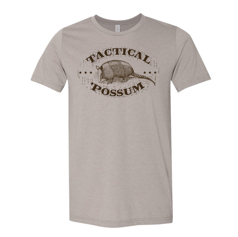Puede incluir: Una camiseta gris claro con un gr&aacute;fico marr&oacute;n de un armadillo y el texto "Tactical Possum".