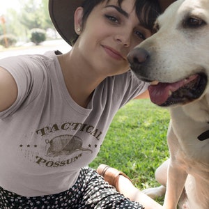 May include: A woman wearing a light gray t-shirt with a brown graphic of an armadillo and the text "Tactical Possum". She is smiling and looking at the camera. A light brown dog is sitting next to her.