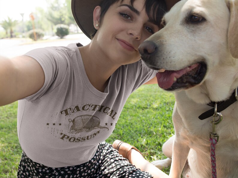 May include: A woman wearing a light gray t-shirt with a brown graphic of an armadillo and the text "Tactical Possum". She is smiling and looking at the camera. A light brown dog is sitting next to her.
