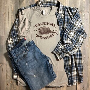 May include: A light gray t-shirt with a brown graphic of an armadillo and the text "Tactical Possum". The t-shirt is paired with blue jeans and a blue and white plaid shirt.