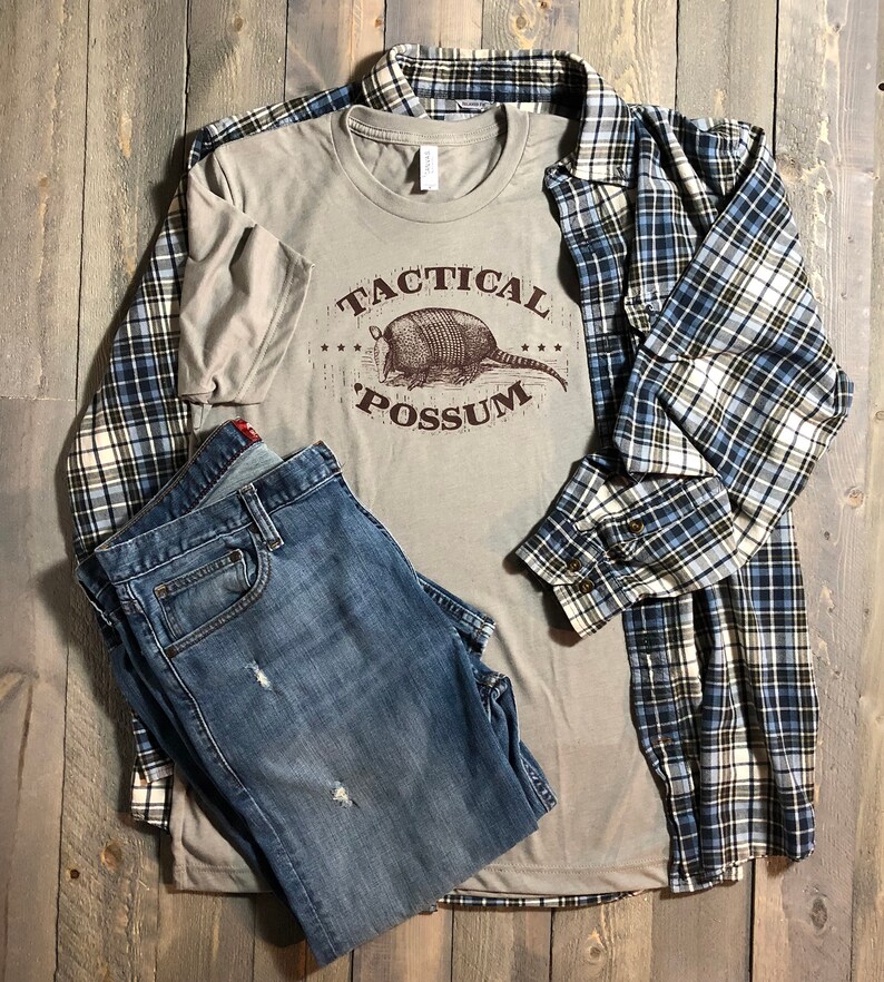 May include: A light gray t-shirt with a brown graphic of an armadillo and the text "Tactical Possum". The t-shirt is paired with blue jeans and a blue and white plaid shirt.
