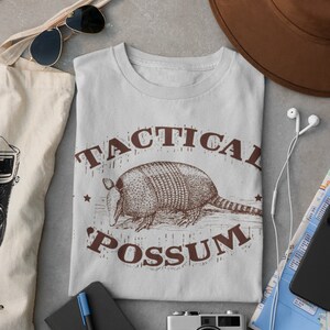 May include: A light gray t-shirt with a brown graphic of an armadillo and the text "Tactical Possum".
