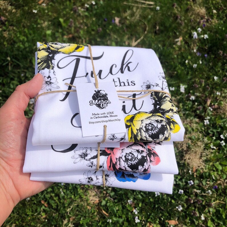 May include: Set of three white tea towels with floral designs and the text "F*ck this sh*t" in black. The towels are tied together with twine and a small tag that reads "Merch Op" and includes information about the shop.