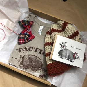 May include: A gift box filled with a brown t-shirt with the text "TACTIC" and a graphic of an armadillo, a red and black plaid bow tie, a brown and white striped knit hat, and a card with the text "Have a Possum Holiday!" and a black and white illustration of a possum.